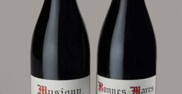 Two wine bottles side by side with red capsule tops and white Grand Cru labels showing vintages 2014 and 2018 (Musigny and Bonnes-Mares).