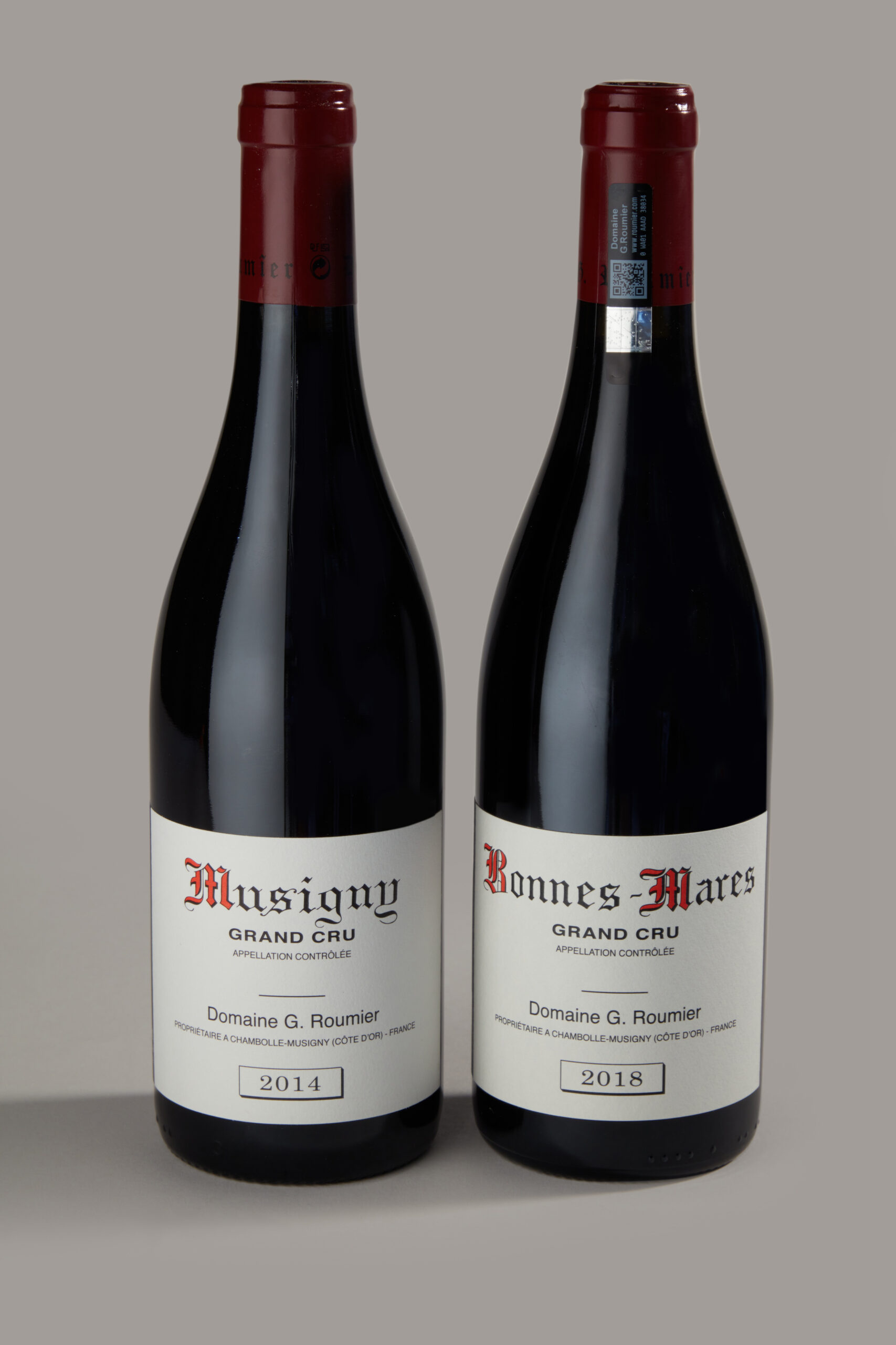 Two wine bottles side by side with red capsule tops and white Grand Cru labels showing vintages 2014 and 2018 (Musigny and Bonnes-Mares).