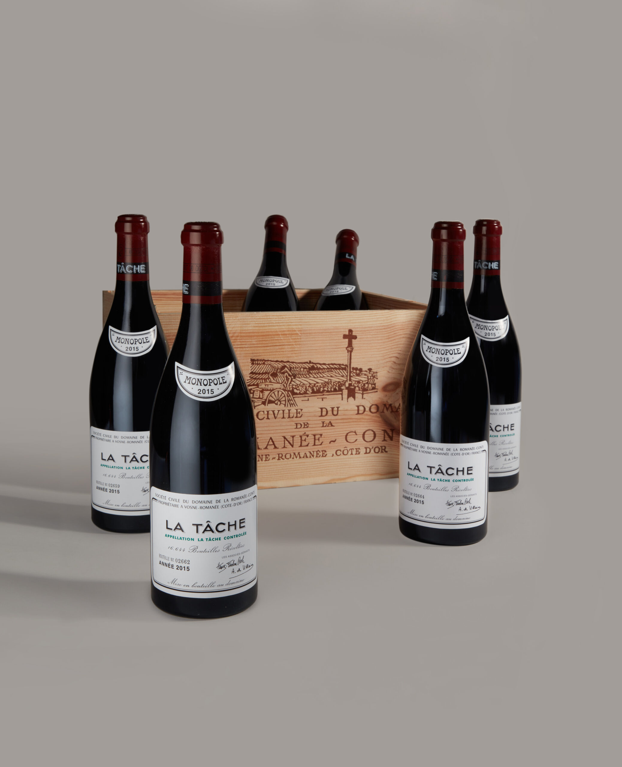 Six bottles of red wine labeled La Tâche arranged around a wooden wine box on a gray background, front bottle centered.
