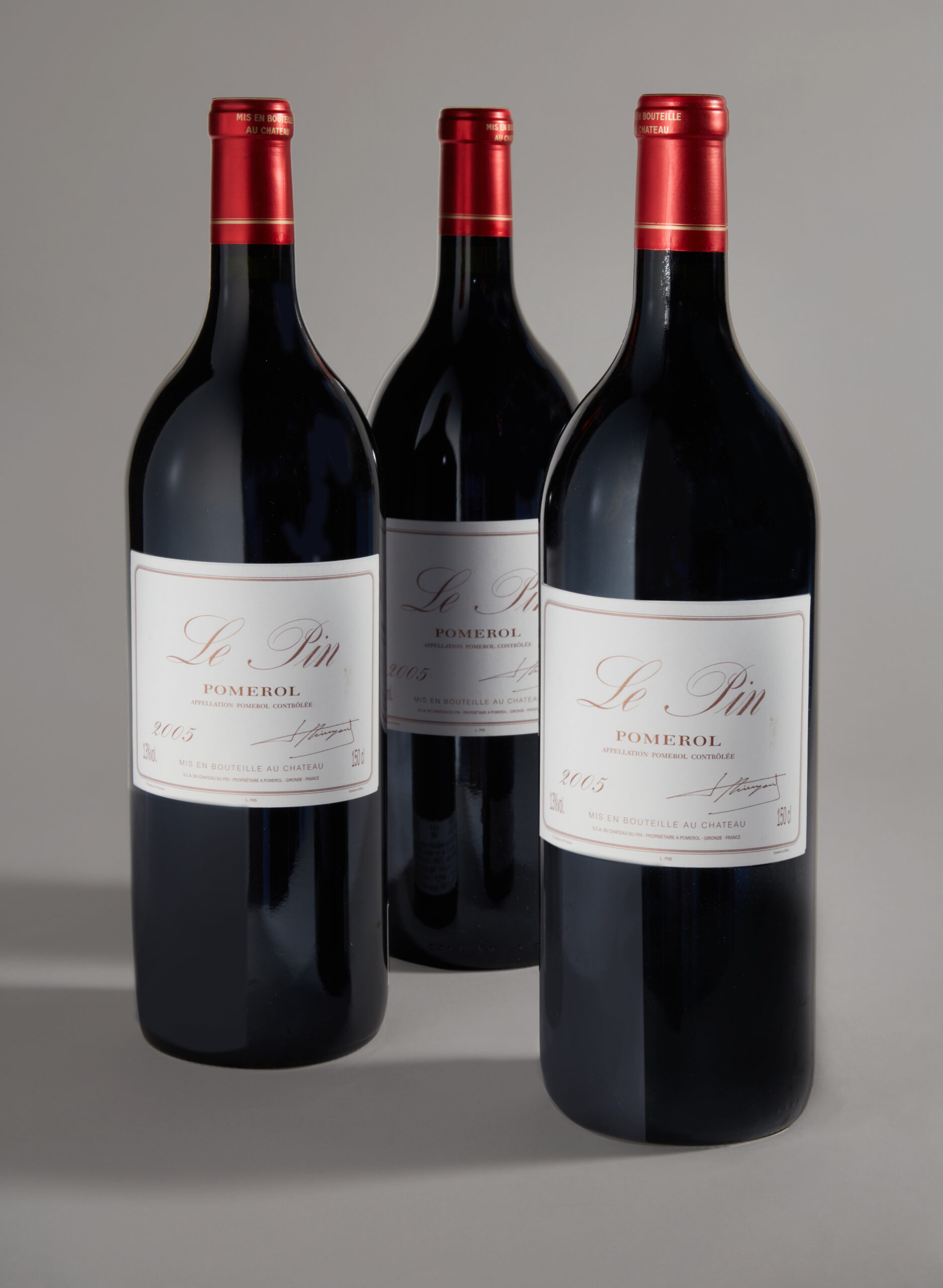 Three dark wine bottles with red foil caps and white labels standing upright against a neutral gray backdrop (Pomerol wine bottles).555